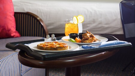 On a table in a stateroom, a tray with a personal size pepperoni pizza, chicken wings and 2 drinks