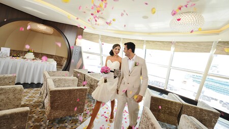 A bridesmaid holding a bouquet walks down a wedding aisle on a ship deck with a groomsman