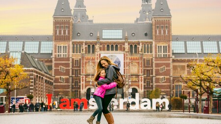 A mother and daughter hugging in front of a reflecting pool and a crowded building facade with a sign that reads I Amsterdam