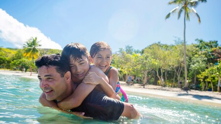 A father wades the shallow waters of a palm fringed beach with 2 children on his back