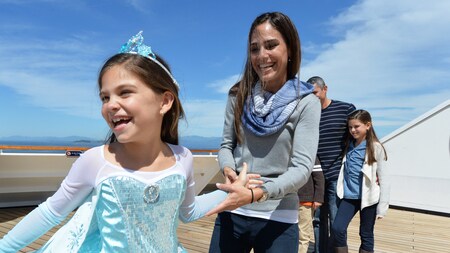 A laughing young girl in a princess dress takes her mother’s hand aboard the deck of a cruise ship