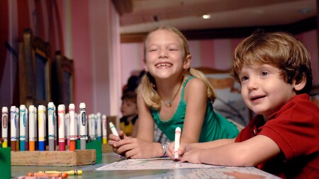 A girl and her younger brother color activity sheets at a table filled with crayons and markers