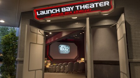 A screen with a logo reading Star Wars Launch Bay and some of the seats of the theater are framed by open side doors above which is a sign with the words Launch Bay Theatre and letter like characters reminiscent of an alien alphabet