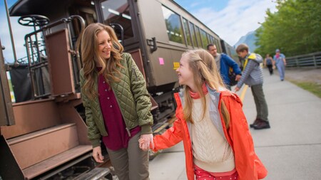 A woman and a girl holding hands and smiling at each other beside a train car