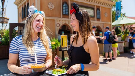 Two women in Minnie Mouse ears eat outside at a standing table