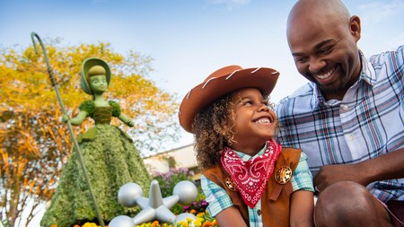 A child dressed like a sheriff and their dad smile next to a topiary of Little Bo Peep