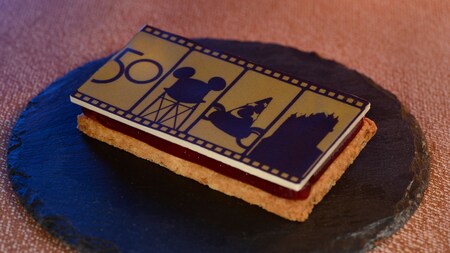 Um sanduíche de cookie decorado com um design dourado destacando o número 50, ao lado de silhuetas de símbolos do Disney’s Hollywood Studios