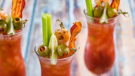 Three glasses of Bloody Marys garnished with bacon, celery, lime, olives, and shrimp 