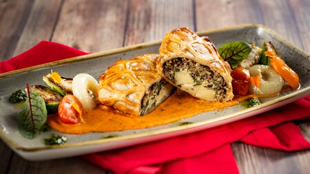 A plate of Vegetable Wellington sliced open on a cream sauce with roasted vegetables and pearl onions  