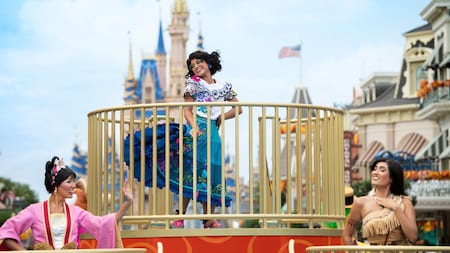 Mirabel stands on a parade float in Magic Kingdom park, with Mulan and Pocahontas alongside her