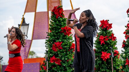 Two women sing and dance on a stage filled with Christmas trees that are decorated with poinsettias