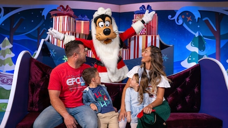 A family of 4 laughing with Goofy dressed in holiday attire in front of a festive backdrop