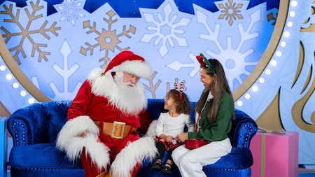 Santa Claus sitting on a sofa with a young girl and her mother in front of a festive backdrop