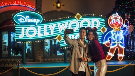 Un couple prend un égoportrait devant une enseigne éclairée indiquant « Disney Jollywood Nights »