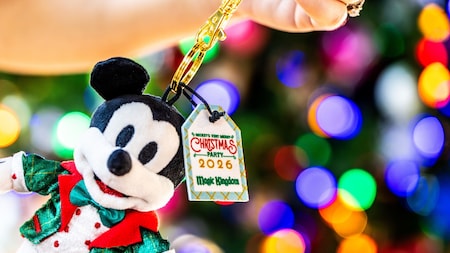 Mickey Mouse holiday plush keychain in festive red and green attire, featuring a visible “2026 Mickey’s Very Merry Christmas Party” key charm, with colorful blurred holiday lights in the background.