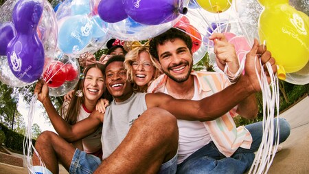 A group of five friends sit closely together, smiling joyfully, surrounded by Mickey Mouse shaped balloons