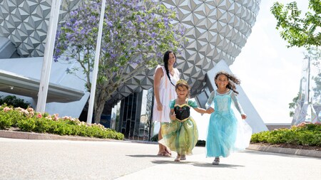 Two young girls running in Anna and Elsa Disney Princess gowns as their mother looks on in front of Spaceship Earth at Epcot