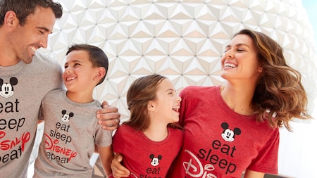 A mother, father and their 2 children sit near Spaceship Earth, wearing T-shirts with text that reads ‘eat, sleep, Disney, repeat’
