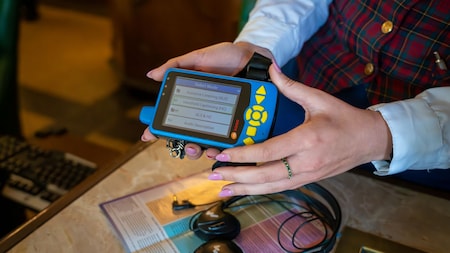 A Cast Member holds the Disney Handheld Device with a pair of headphones and a map on the counter.