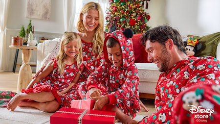 A family of 4 opening gifts while wearing matching holiday themed pajamas featuring Mickey Mouse wearing a Santa hat