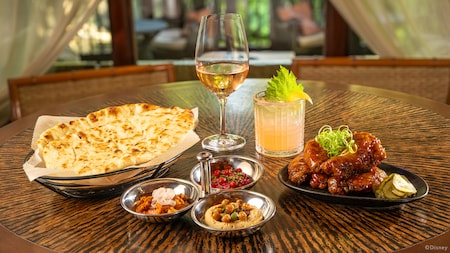 A table including a basket of naan bread, a trio of dips and a plate of wings next to a glass of wine and a cocktail
