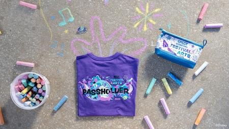 A shirt with text that says, ‘Epcot International Festival of the Arts 2026 Passholder’ placed on top of sidewalk art next to pieces of chalk 