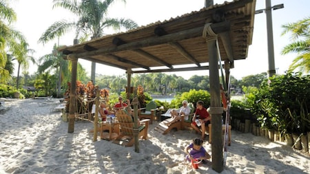 A family of 6 seated in Adirondack chairs under a wooden gazebo like structure surrounded by palm trees at Disney’s Typhoon Lagoon water park