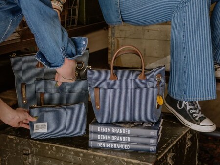 Canvas and leather tote bags and pouches displayed on a vintage metal trunk stacked with books titled ‘Denim Branded,’ with 2 people seated nearby