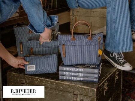 Canvas and leather tote bags and pouches displayed on a vintage metal trunk stacked with books titled ‘Denim Branded,’ with 2 people seated nearby