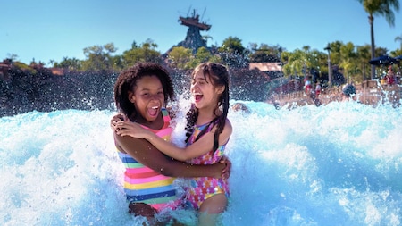2 girls standing in a wave pool, holding one another while a wave crashes behind them