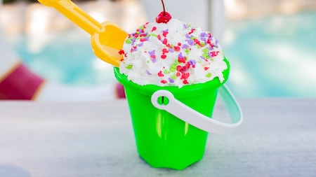 A sundae with whipped cream, sprinkles and a cherry served in a sand pail with a small sand shovel