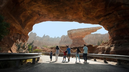 A group of 6 Guests holding hands as they walk along a road under a canyon tunnel at Cars Land