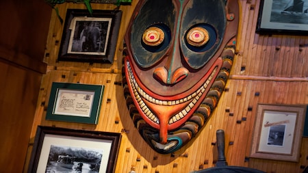 A colorful carved tiki mask displayed on a bamboo wall alongside vintage framed photos at Trader Sam's Enchanted Tiki Bar