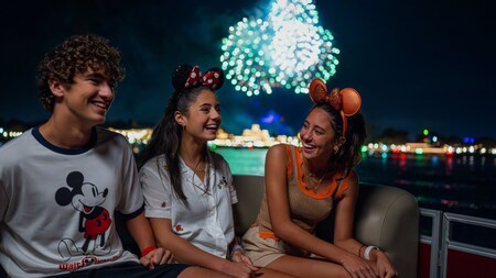 Three teens smiling on a fireworks cruise as fireworks burst above Seven Seas Lagoon