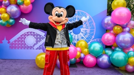 Mickey Mouse standing with arms outstretched near a photo op wall with balloons