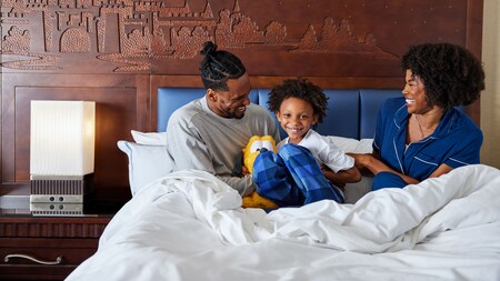 A father, mother and their young son relaxing on a hotel bed while the son holds a Pluto plush toy