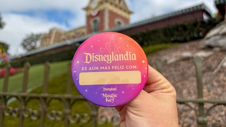 A hand holding a button with Spanish text reading, “Disneyland is even happier with,” and a blank space for the Guest's name