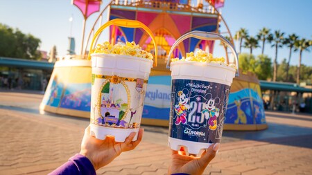 Hands holding a Disneyland Resort 70th Celebration popcorn bucket and a Disney California Adventure Park 25th Anniversary popcorn bucket.