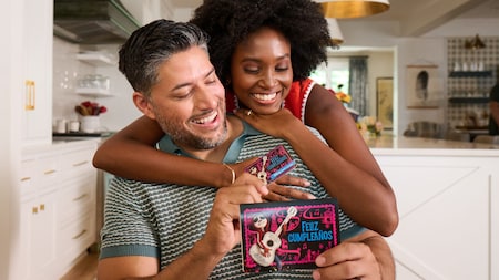 A smiling couple embracing while holding a Coco themed Feliz Cumpleaños gift card and a greeting card