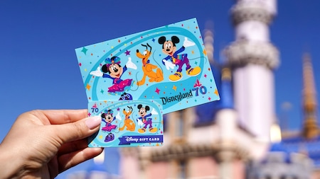 Una mano sosteniendo una tarjeta de regalo de la celebración del 70.º aniversario de Disneyland Resort frente al Sleeping Beauty Castle