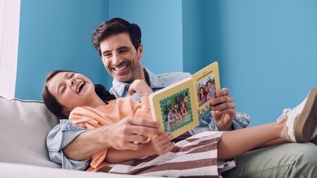 A father and child laugh on a couch while looking at a family photo album
