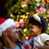 A man in a Santa hat stands with his child in front of a Christmas tree