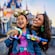 A girl wearing a Disneyland 70th anniversary ear headband and her mother, both playing with a bubble blower near Sleeping Beauty Castle