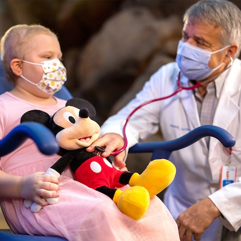 Un médico con una mascarilla arrodilló junto a un niño sentado en una silla de ruedas con un peluche de Mickey mouse
