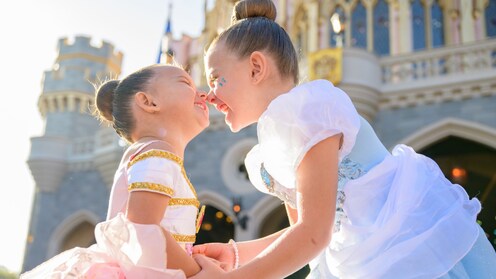 Two girls dressed as Belle and Cinderella touching noses in front of Sleeping Beauty Castle