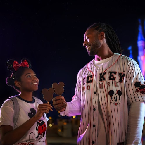 Two smiling young adults wearing Mickey Mouse apparel and holding Mickey Mouse ice cream bars in front of Cinderella Castle