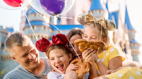 Une famille souriant et s’embrassant les uns les autres tout en tenant des ballons et des bretzels Mickey Mouse devant le Cinderella Castle