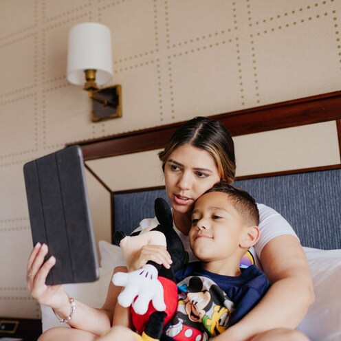 A woman sits on a bed in a hotel room and shows an I Pad screen to her son, who sits with her