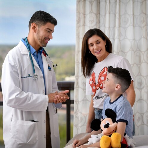 A doctor talking to a boy sitting on a bed with a Mickey Mouse plush while his mother stands next to him