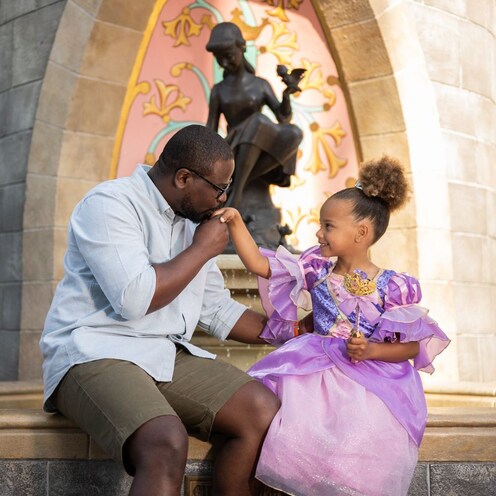 A father kissing the hand of his young daughter who is wearing a princess gown in front of Cinderella Fountain